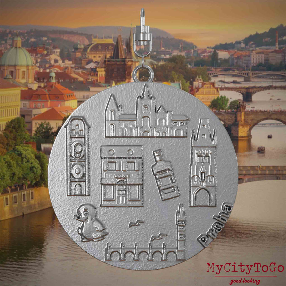 Halskette mit Drehwirbel und Vorderseite eines Schmuckanhängers aus Edelstahl, auf dem Reliefs bekannter Sehenswürdigkeiten der Stadt Prag abgebildet sind. Den Hintergrund bildet eine Ansicht der Stadt Prag.
