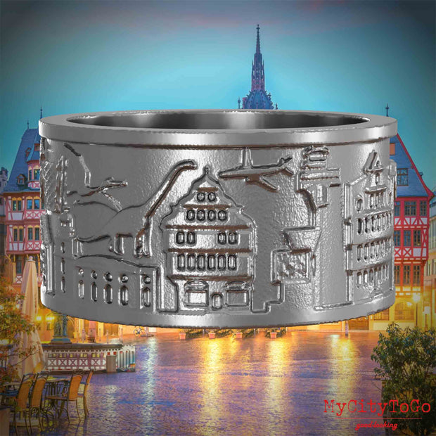 Ring aus Edelstahl mit Reliefs bekannter Sehenswürdigkeiten der Stadt Frankfurt. Den Hintergrund bildet eine Ansicht der Stadt Frankfurt.