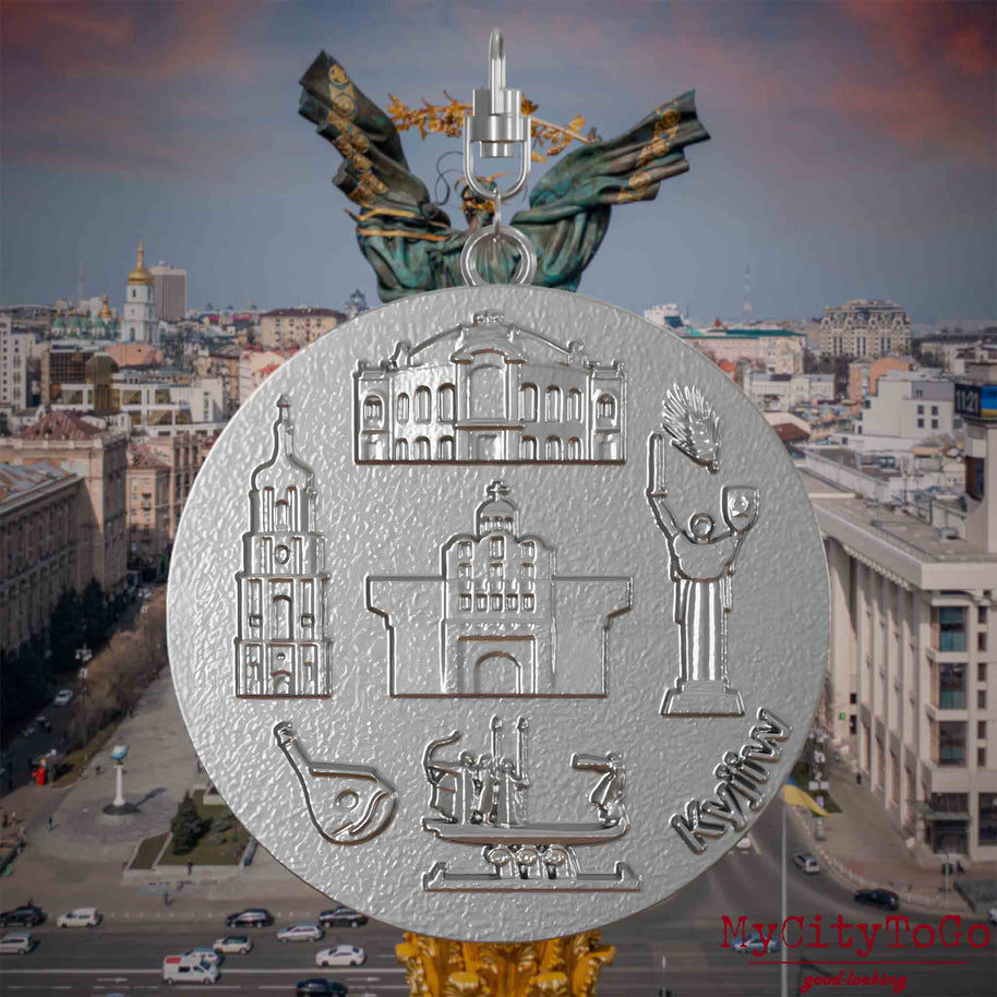 Halskette mit Drehwirbel und Vorderseite eines Schmuckanhängers aus Edelstahl, auf dem Reliefs bekannter Sehenswürdigkeiten der Stadt Kyjiw abgebildet sind. Den Hintergrund bildet eine Ansicht der Stadt Kyjiw.
