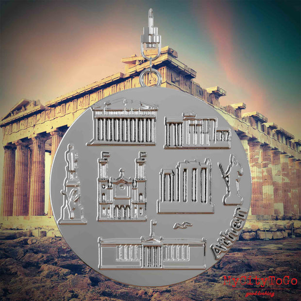 Halskette mit Drehwirbel und Vorderseite eines silbernen Schmuckanhängers, auf dem Reliefs bekannter Sehenswürdigkeiten der Stadt Athen abgebildet sind. Den Hintergrund bildet eine Ansicht der Stadt Athen.
