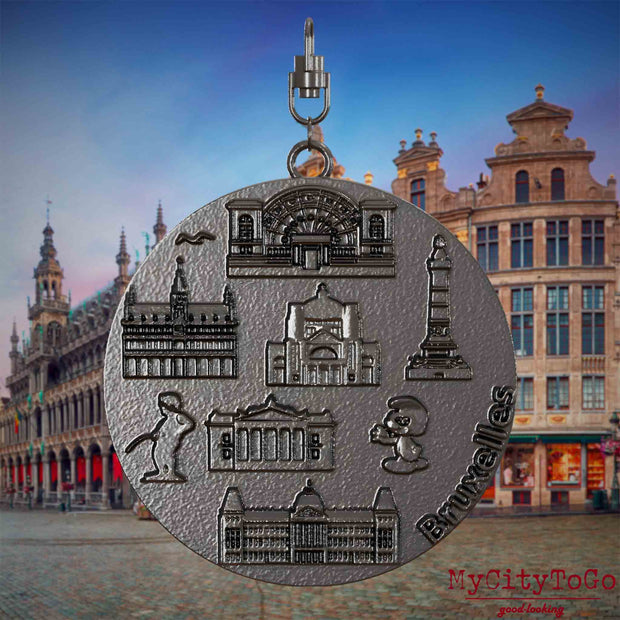 Halskette mit Drehwirbel und Vorderseite eines Schmuckanhängers aus Titan, auf dem Reliefs bekannter Sehenswürdigkeiten der Stadt Bruessel abgebildet sind. Den Hintergrund bildet eine Ansicht der Stadt Bruessel.
