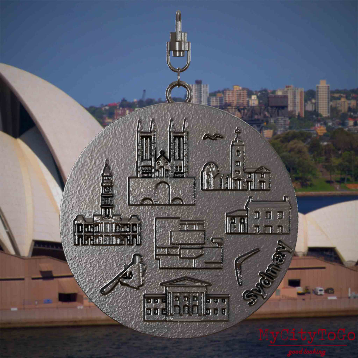 Halskette mit Drehwirbel und Rückseite eines Schmuckanhängers aus Titan, auf dem Reliefs bekannter Sehenswürdigkeiten der Stadt Sydney abgebildet sind. Den Hintergrund bildet eine Ansicht der Stadt Sydney.
