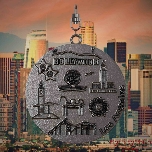 Halskette mit Drehwirbel und Vorderseite eines Schmuckanhängers aus Titan, auf dem Reliefs bekannter Sehenswürdigkeiten der Stadt Los Angeles abgebildet sind. Den Hintergrund bildet eine Ansicht der Stadt Los Angeles.
