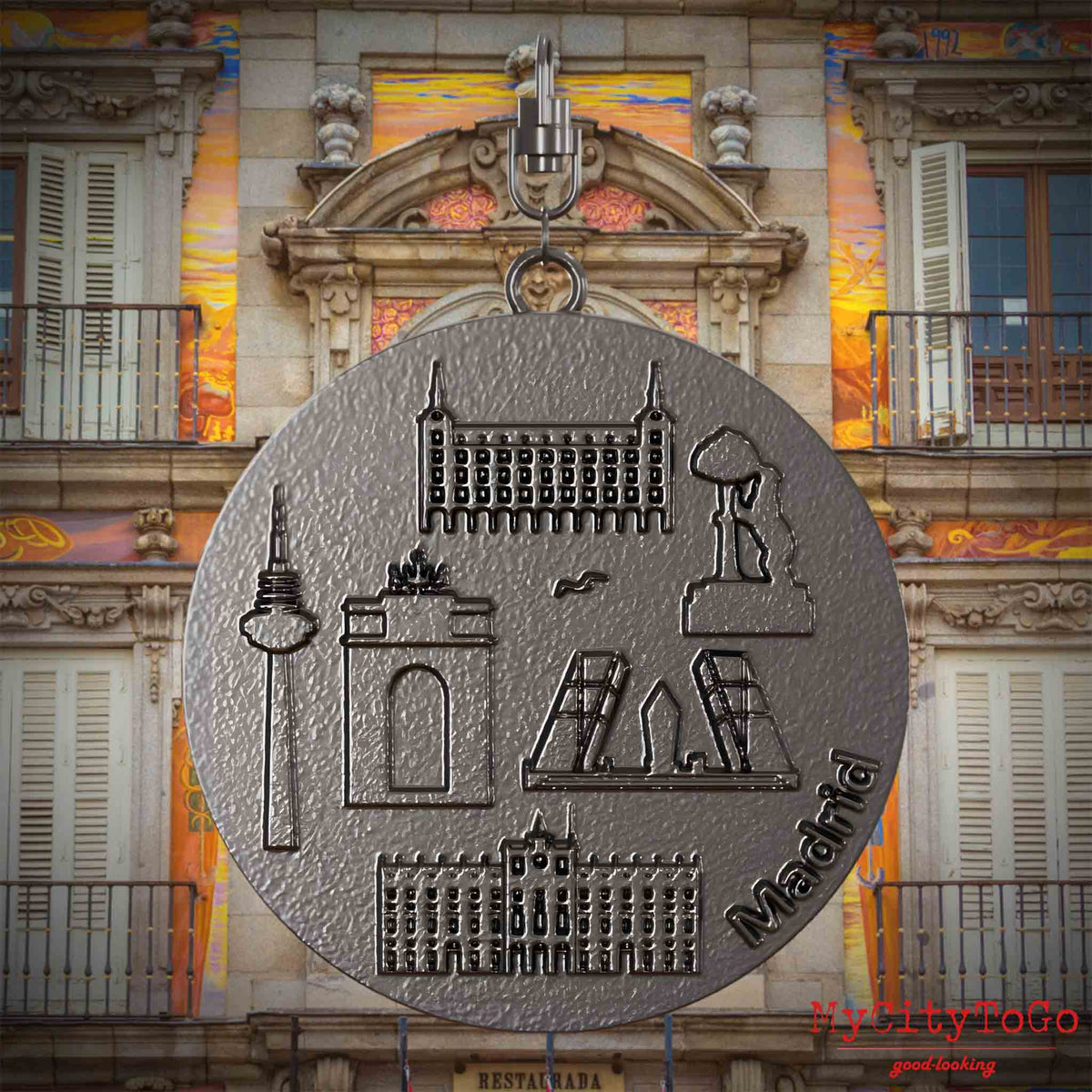 Halskette mit Drehwirbel und Vorderseite eines Schmuckanhängers aus Titan, auf dem Reliefs bekannter Sehenswürdigkeiten der Stadt Madrid abgebildet sind. Den Hintergrund bildet eine Ansicht der Stadt Madrid.
