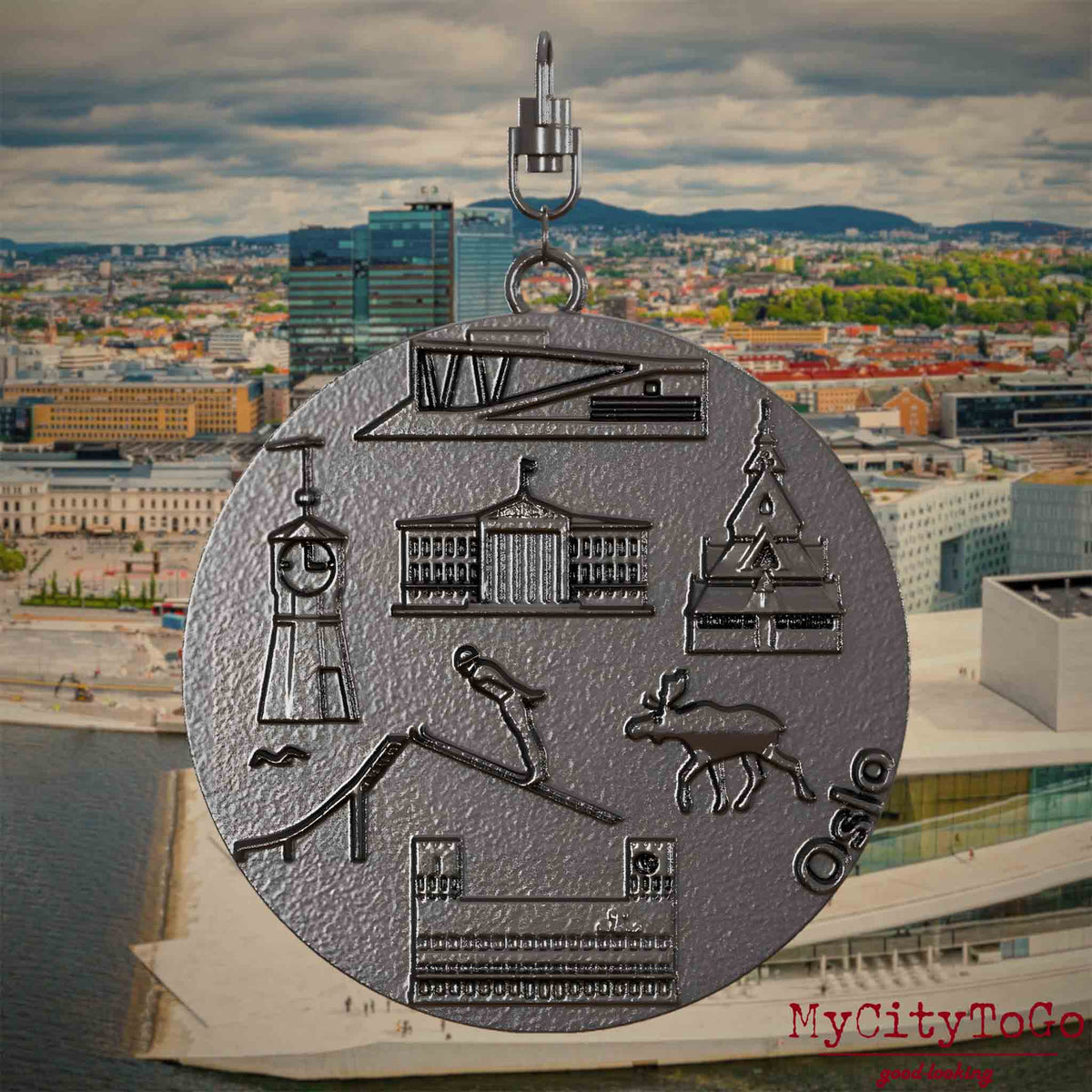Halskette mit Drehwirbel und Vorderseite eines Schmuckanhängers aus Titan, auf dem Reliefs bekannter Sehenswürdigkeiten der Stadt Oslo abgebildet sind. Den Hintergrund bildet eine Ansicht der Stadt Oslo.
