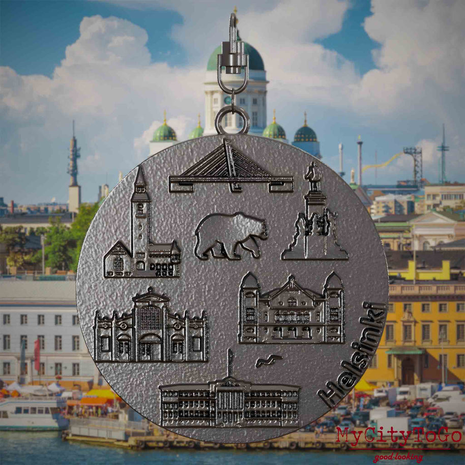 Halskette mit Drehwirbel und Rückseite eines Schmuckanhängers aus Titan, auf dem Reliefs bekannter Sehenswürdigkeiten der Stadt Helsinki abgebildet sind. Den Hintergrund bildet eine Ansicht der Stadt Helsinki.
