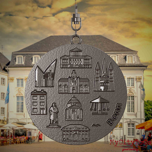 Halskette mit Drehwirbel und Vorderseite eines Schmuckanhängers aus Titan, auf dem Reliefs bekannter Sehenswürdigkeiten der Stadt Bonn abgebildet sind. Den Hintergrund bildet eine Ansicht der Stadt Bonn.
