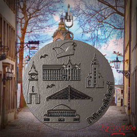 Halskette mit Drehwirbel und Rückseite eines Schmuckanhängers aus Titan, auf dem Reliefs bekannter Sehenswürdigkeiten der Stadt Duesseldorf abgebildet sind. Den Hintergrund bildet eine Ansicht der Stadt Duesseldorf.