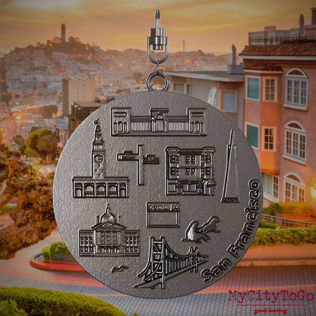 Halskette mit Drehwirbel und Vorderseite eines Schmuckanhängers aus Titan, auf dem Reliefs bekannter Sehenswürdigkeiten der Stadt San Francisco abgebildet sind. Den Hintergrund bildet eine Ansicht der Stadt San Francisco.
