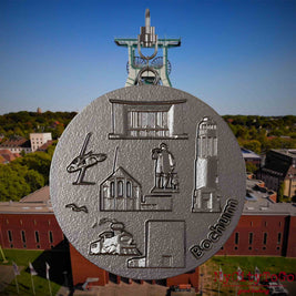 Halskette mit Drehwirbel und Rückseite eines Schmuckanhängers aus Titan, auf dem Reliefs bekannter Sehenswürdigkeiten der Stadt Bochum abgebildet sind. Den Hintergrund bildet eine Ansicht der Stadt Bochum .
