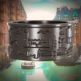 Ring aus Titan mit Reliefs bekannter Sehenswürdigkeiten der Stadt Hamburg. Den Hintergrund bildet eine Ansicht der Stadt Hamburg.
