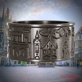 Ring aus Titan mit Reliefs bekannter Sehenswürdigkeiten der Stadt Muenchen. Den Hintergrund bildet eine Ansicht der Stadt Muenchen.