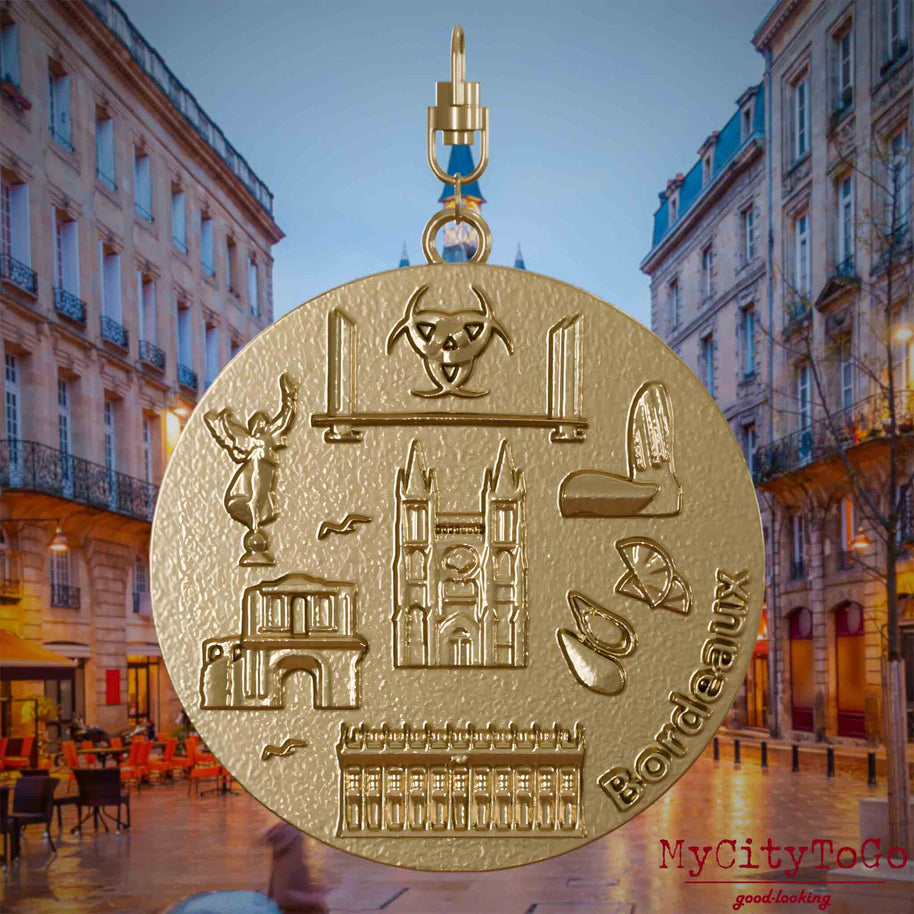 Halskette mit Drehwirbel und Vorderseite eines Schmuckanhängers aus vergoldetem Edelstahl, auf dem Reliefs bekannter Sehenswürdigkeiten der Stadt Bordeaux abgebildet sind. Den Hintergrund bildet eine Ansicht der Stadt Bordeaux.
