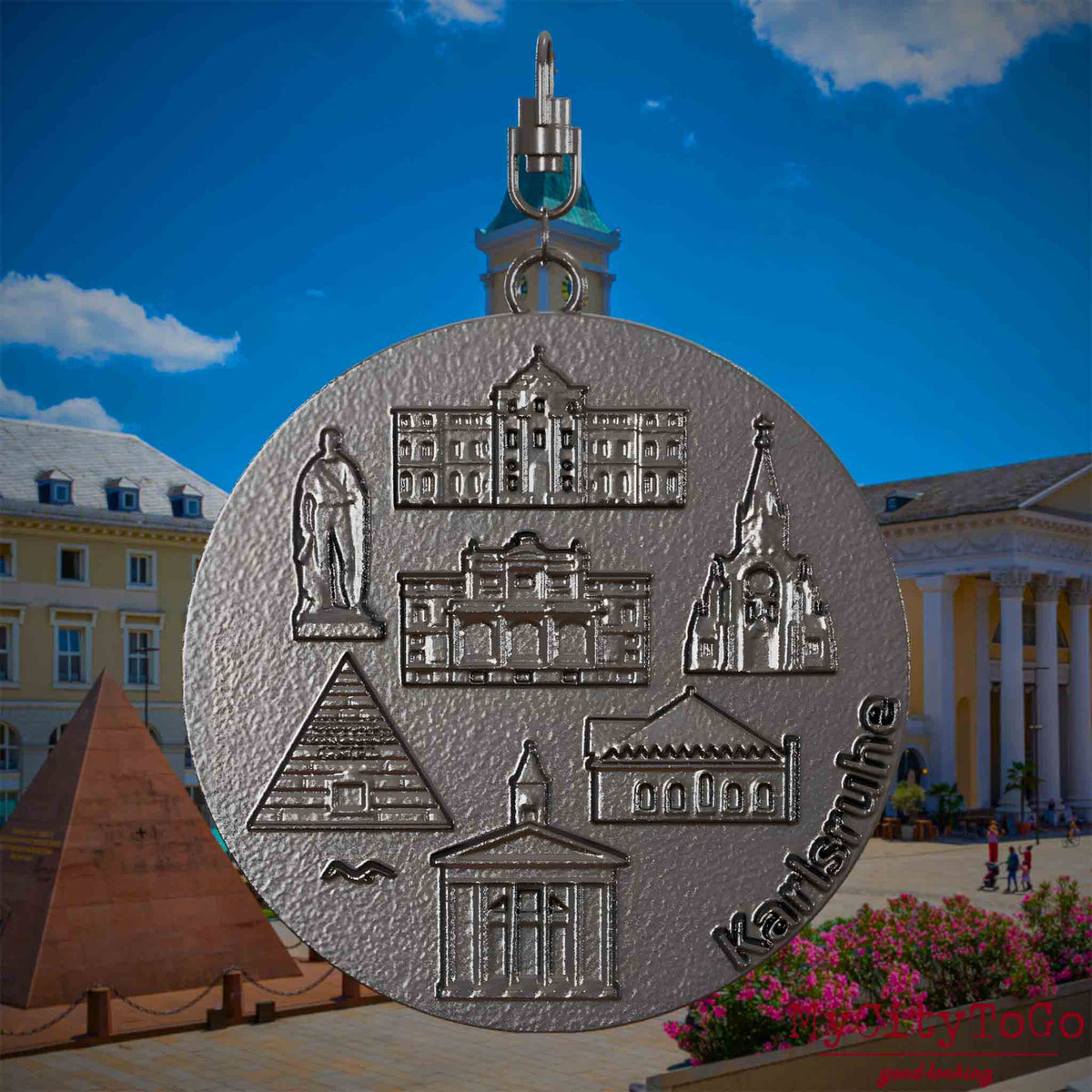 Halskette mit Drehwirbel und Vorderseite eines Schmuckanhängers aus Titan, auf dem Reliefs bekannter Sehenswürdigkeiten der Stadt Karlsruhe abgebildet sind. Den Hintergrund bildet eine Ansicht der Stadt Karlsruhe .
