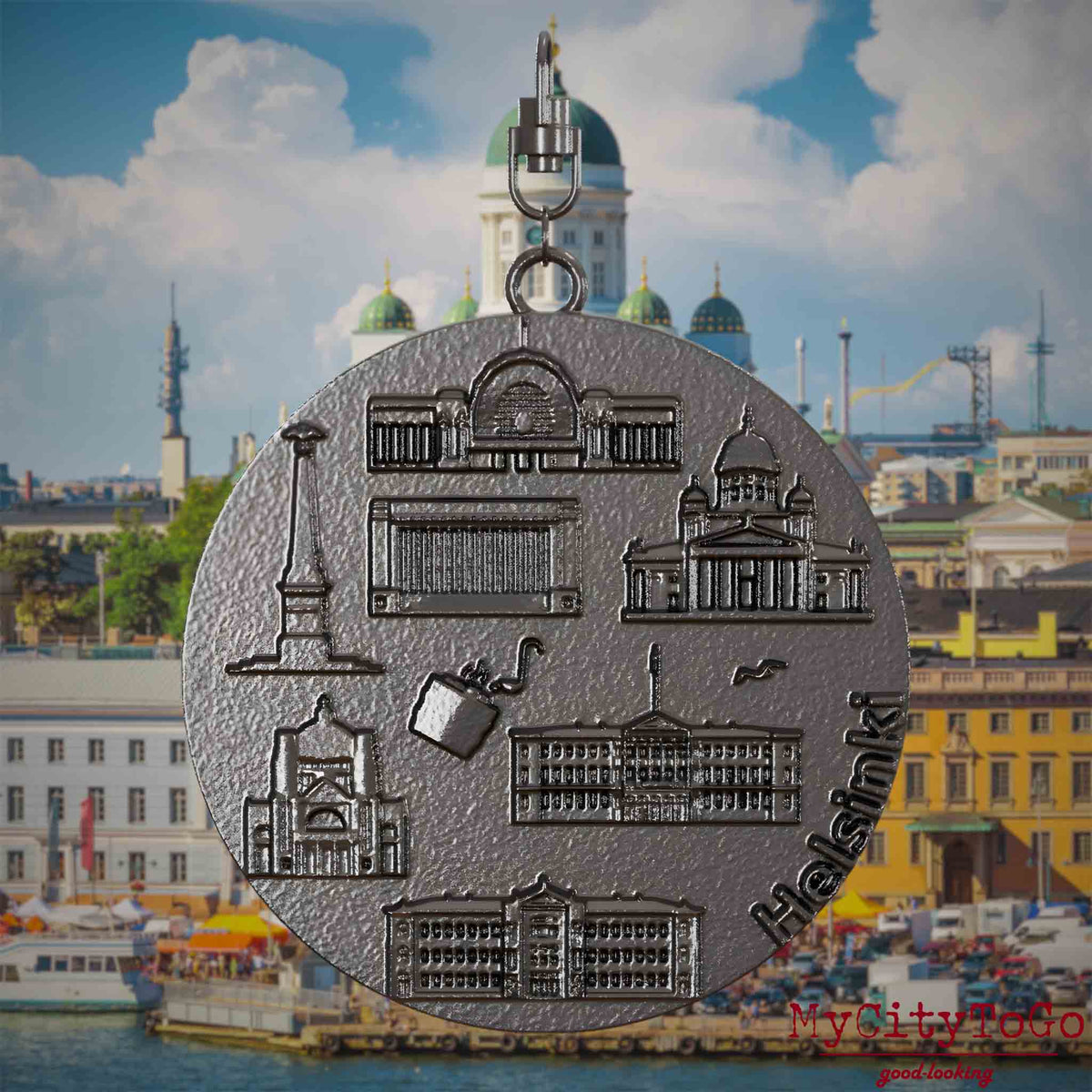 Halskette mit Drehwirbel und Vorderseite eines Schmuckanhängers aus Titan, auf dem Reliefs bekannter Sehenswürdigkeiten der Stadt Helsinki abgebildet sind. Den Hintergrund bildet eine Ansicht der Stadt Helsinki.
