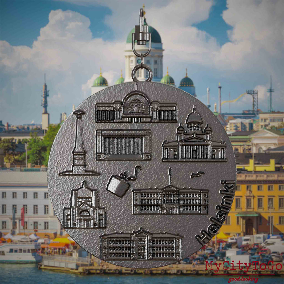 Halskette mit Drehwirbel und Vorderseite eines Schmuckanhängers aus Titan, auf dem Reliefs bekannter Sehenswürdigkeiten der Stadt Helsinki abgebildet sind. Den Hintergrund bildet eine Ansicht der Stadt Helsinki.
