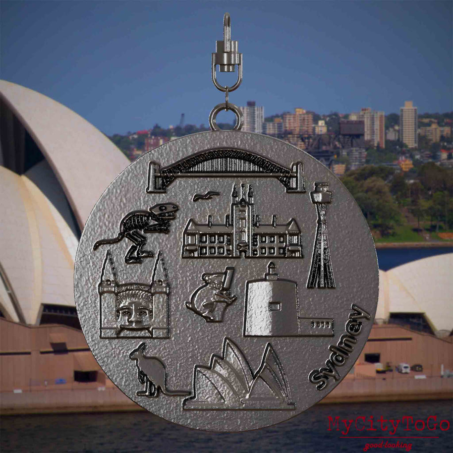 Halskette mit Drehwirbel und Vorderseite eines Schmuckanhängers aus Titan, auf dem Reliefs bekannter Sehenswürdigkeiten der Stadt Sydney abgebildet sind. Den Hintergrund bildet eine Ansicht der Stadt Sydney.
