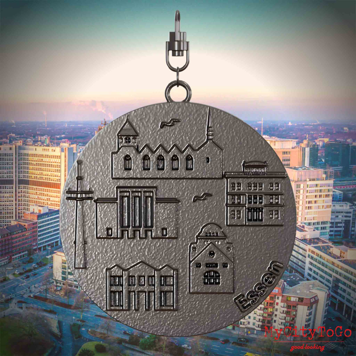 Halskette mit Drehwirbel und Vorderseite eines Schmuckanhängers aus Titan, auf dem Reliefs bekannter Sehenswürdigkeiten der Stadt Essen abgebildet sind. Den Hintergrund bildet eine Ansicht der Stadt Essen.
