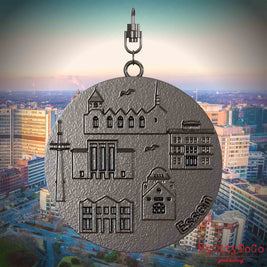 Halskette mit Drehwirbel und Vorderseite eines Schmuckanhängers aus Titan, auf dem Reliefs bekannter Sehenswürdigkeiten der Stadt Essen abgebildet sind. Den Hintergrund bildet eine Ansicht der Stadt Essen.
