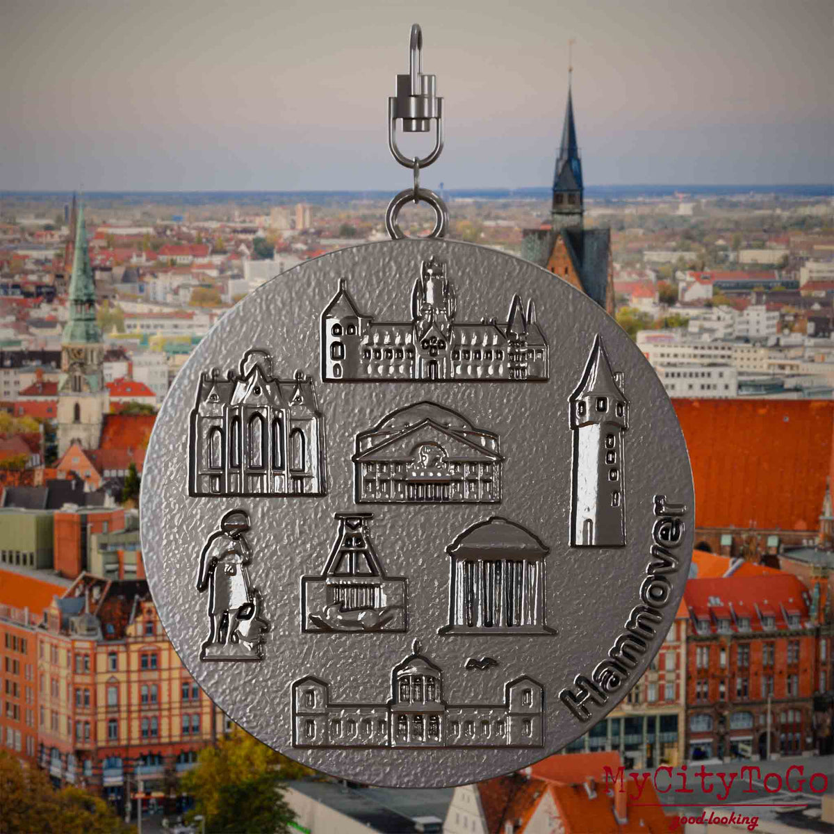 Halskette mit Drehwirbel und Vorderseite eines Schmuckanhängers aus Titan, auf dem Reliefs bekannter Sehenswürdigkeiten der Stadt Hannover abgebildet sind. Den Hintergrund bildet eine Ansicht der Stadt Hannover .
