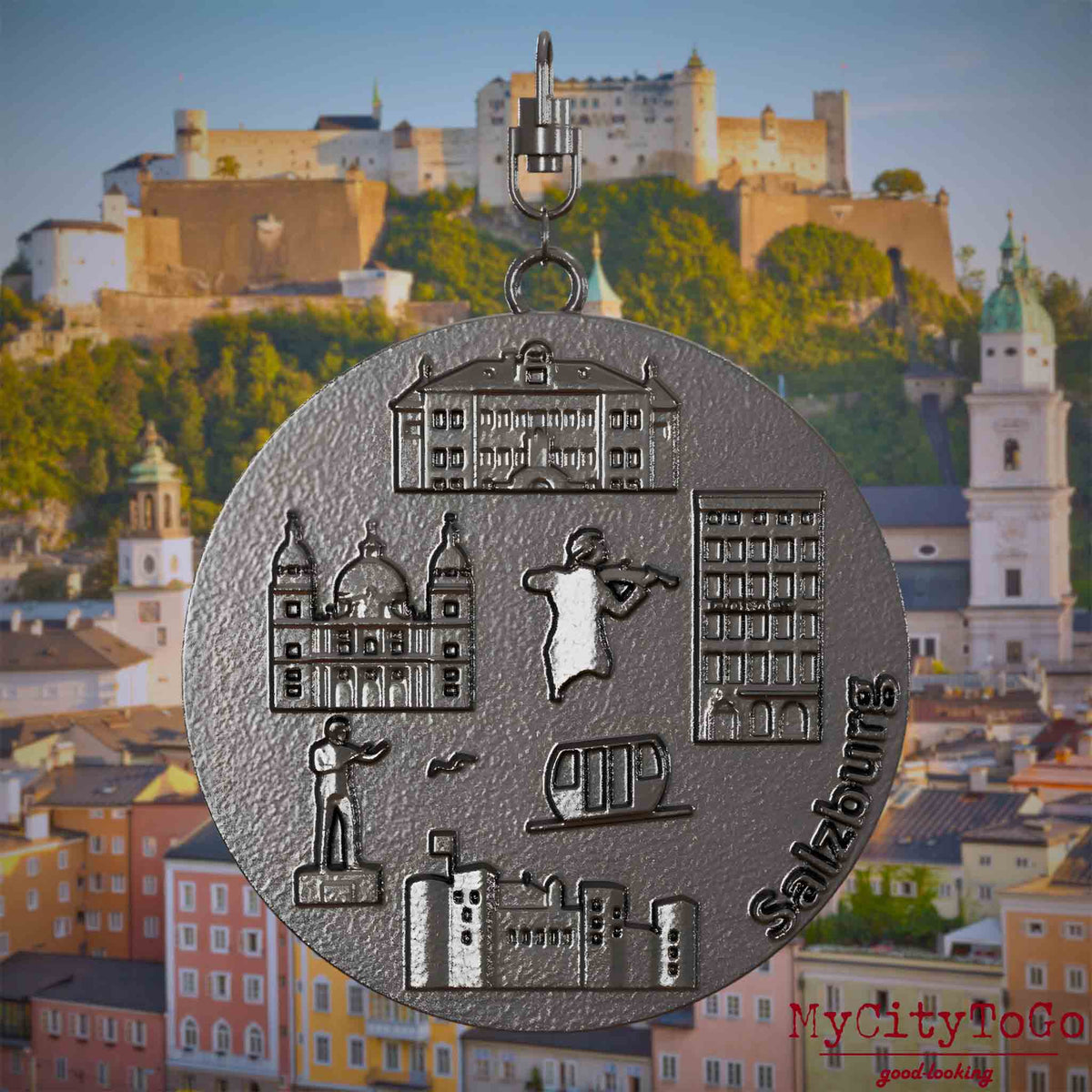 Halskette mit Drehwirbel und Vorderseite eines Schmuckanhängers aus Titan, auf dem Reliefs bekannter Sehenswürdigkeiten der Stadt Salzburg abgebildet sind. Den Hintergrund bildet eine Ansicht der Stadt Salzburg.
