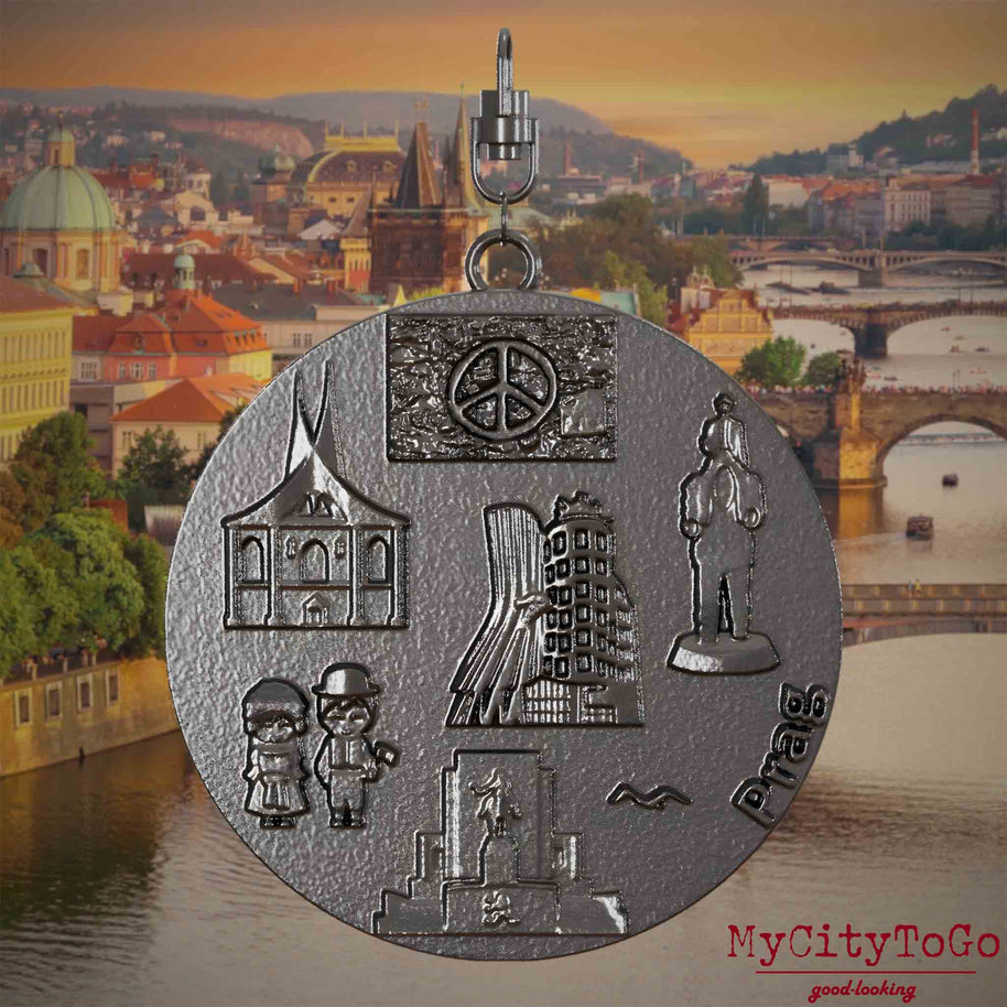 Halskette mit Drehwirbel und Rückseite eines Schmuckanhängers aus Titan, auf dem Reliefs bekannter Sehenswürdigkeiten der Stadt Prag abgebildet sind. Den Hintergrund bildet eine Ansicht der Stadt Prag.

