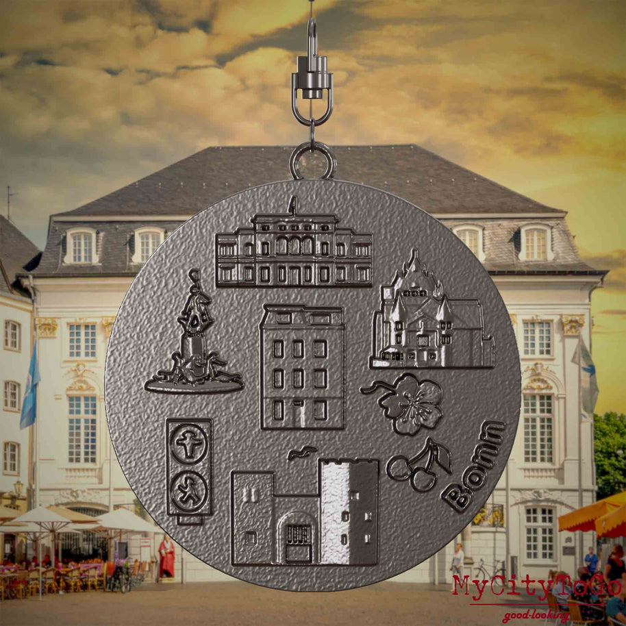 Halskette mit Drehwirbel und Rückseite eines Schmuckanhängers aus Titan, auf dem Reliefs bekannter Sehenswürdigkeiten der Stadt Bonn abgebildet sind. Den Hintergrund bildet eine Ansicht der Stadt Bonn.
