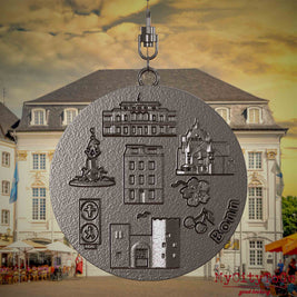 Halskette mit Drehwirbel und Rückseite eines Schmuckanhängers aus Titan, auf dem Reliefs bekannter Sehenswürdigkeiten der Stadt Bonn abgebildet sind. Den Hintergrund bildet eine Ansicht der Stadt Bonn.
