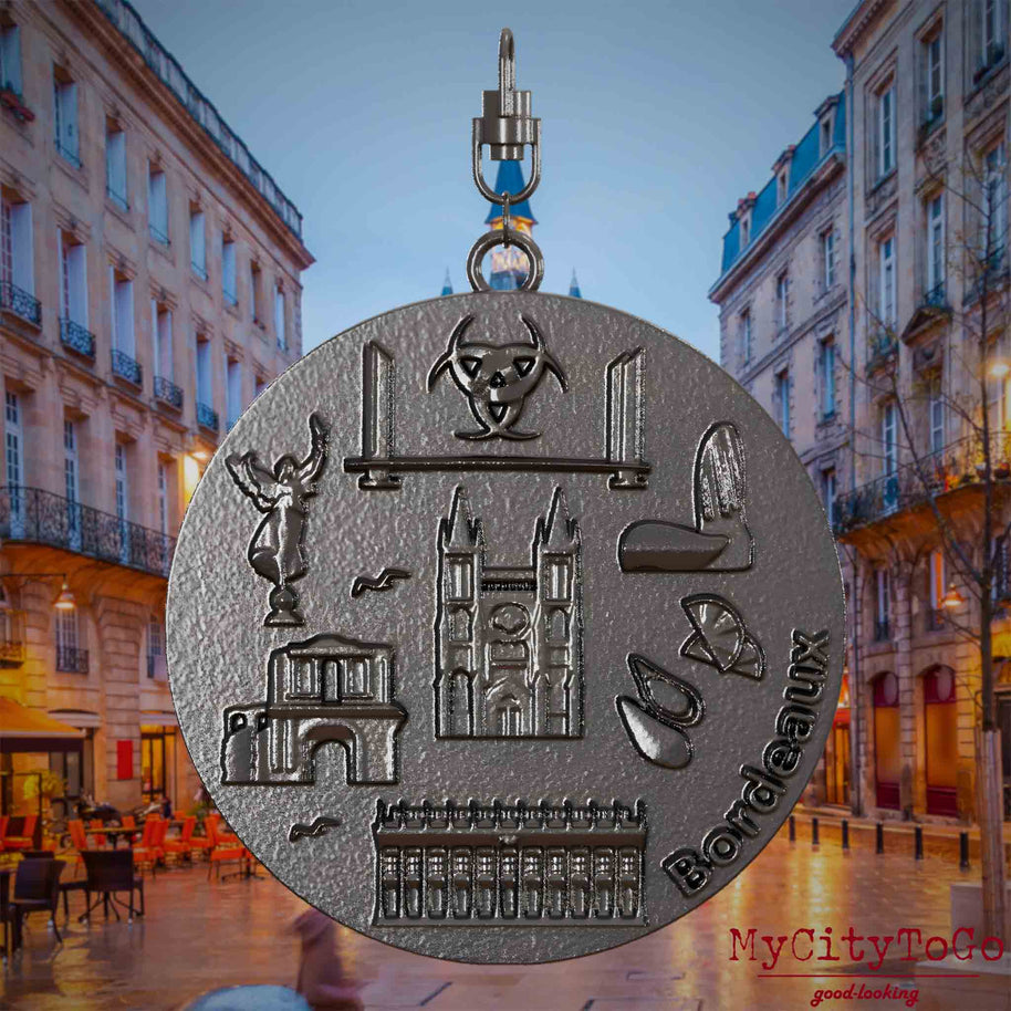 Halskette mit Drehwirbel und Vorderseite eines Schmuckanhängers aus Titan, auf dem Reliefs bekannter Sehenswürdigkeiten der Stadt Bordeaux abgebildet sind. Den Hintergrund bildet eine Ansicht der Stadt Bordeaux.
