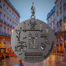 Halskette mit Drehwirbel und Vorderseite eines Schmuckanhängers aus Titan, auf dem Reliefs bekannter Sehenswürdigkeiten der Stadt Bordeaux abgebildet sind. Den Hintergrund bildet eine Ansicht der Stadt Bordeaux.
