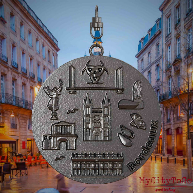 Halskette mit Drehwirbel und Vorderseite eines Schmuckanhängers aus Titan, auf dem Reliefs bekannter Sehenswürdigkeiten der Stadt Bordeaux abgebildet sind. Den Hintergrund bildet eine Ansicht der Stadt Bordeaux.
