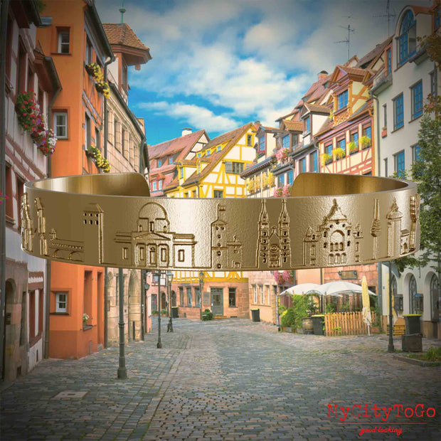 Armreif aus vergoldetem Edelstahl mit Reliefs bekannter Sehenswürdigkeiten der Stadt Nürnberg. Den Hintergrund bildet eine Ansicht der Stadt  Nuernberg.
