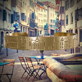 Armreif aus vergoldetem Edelstahl mit Reliefs bekannter Sehenswürdigkeiten der Stadt Zuerich. Den Hintergrund bildet eine Ansicht der Stadt Zuerich.