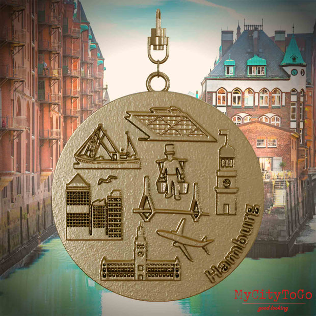 Halskette mit Drehwirbel und Vorderseite eines Schmuckanhängers aus vergoldetem Edelstahl, auf dem Reliefs bekannter Sehenswürdigkeiten der Stadt Hamburg abgebildet sind. Den Hintergrund bildet eine Ansicht der Stadt Hamburg.