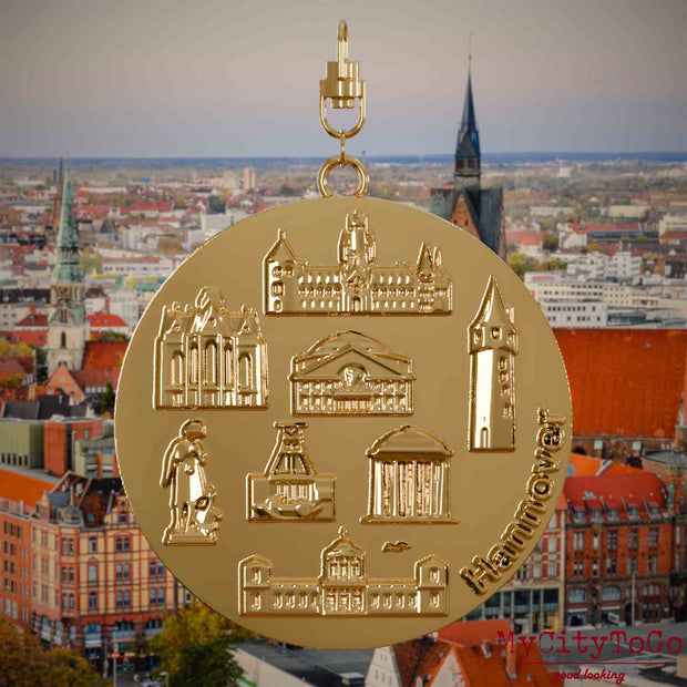 Halskette mit Drehwirbel und Vorderseite eines vergoldeten Schmuckanhängers, auf dem Reliefs bekannter Sehenswürdigkeiten der Stadt Hannover abgebildet sind. Den Hintergrund bildet eine Ansicht der Stadt Hannover .
