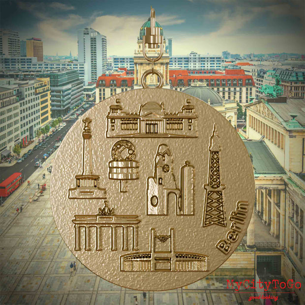 Halskette mit Drehwirbel und Rückseite eines Schmuckanhängers aus vergoldetem Edelstahl, auf dem Reliefs bekannter Sehenswürdigkeiten der Stadt Berlin abgebildet sind. Den Hintergrund bildet eine Ansicht der Stadt Berlin.