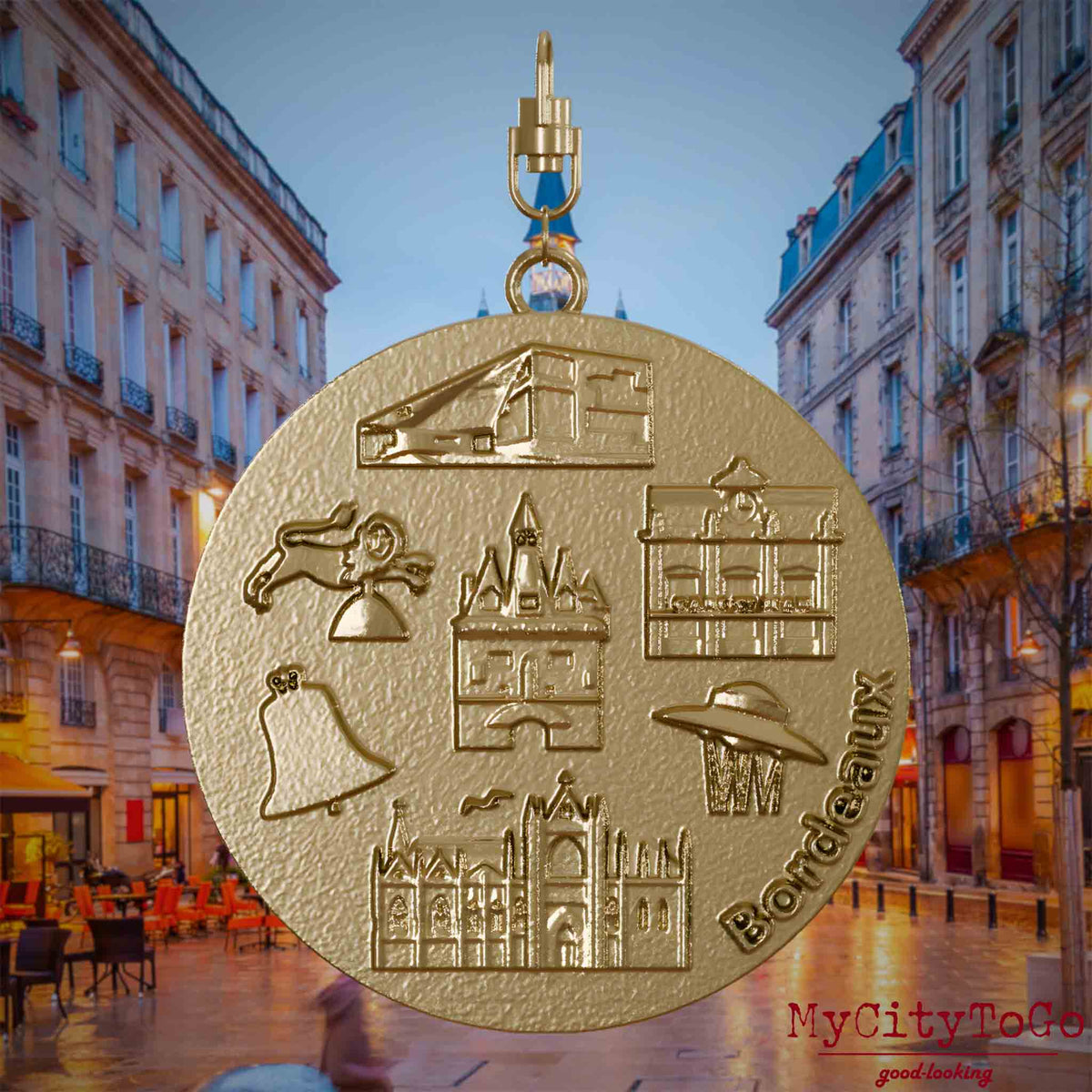 Halskette mit Drehwirbel und Rückseite eines Schmuckanhängers aus vergoldetem Edelstahl, auf dem Reliefs bekannter Sehenswürdigkeiten der Stadt Bordeaux abgebildet sind. Den Hintergrund bildet eine Ansicht der Stadt Bordeaux.
