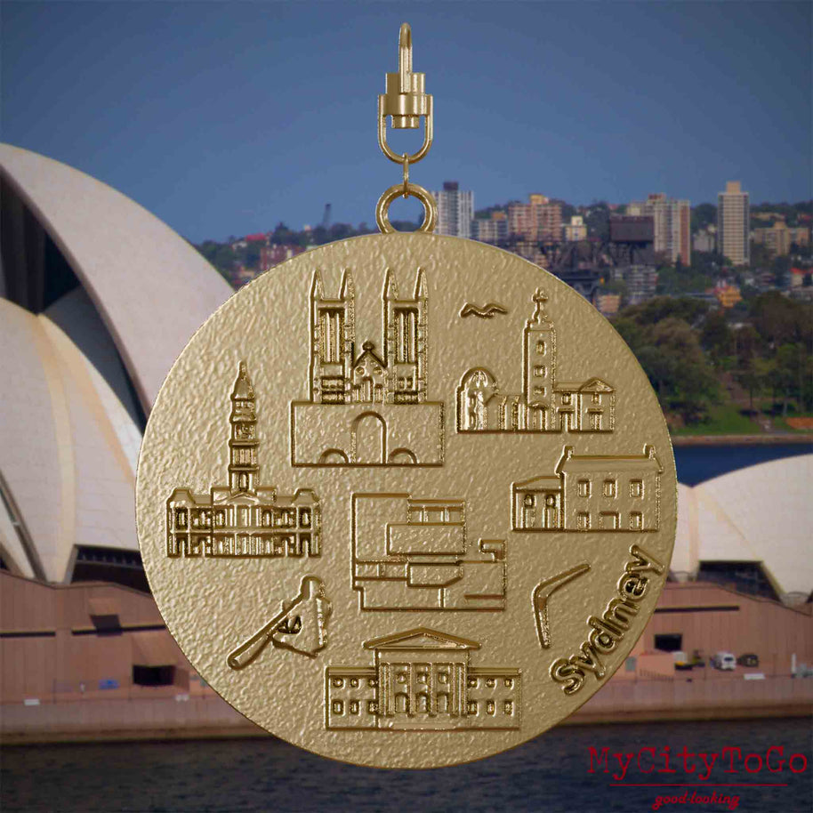 Halskette mit Drehwirbel und Rückseite eines Schmuckanhängers aus vergoldetem Edelstahl, auf dem Reliefs bekannter Sehenswürdigkeiten der Stadt Sydney abgebildet sind. Den Hintergrund bildet eine Ansicht der Stadt Sydney.

