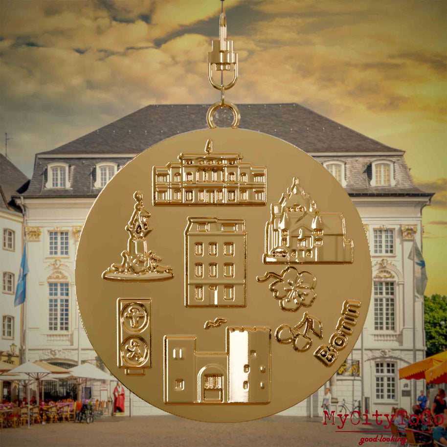 Halskette mit Drehwirbel und Rückseite eines vergoldeten Schmuckanhängers, auf dem Reliefs bekannter Sehenswürdigkeiten der Stadt Bonn abgebildet sind. Den Hintergrund bildet eine Ansicht der Stadt Bonn.
