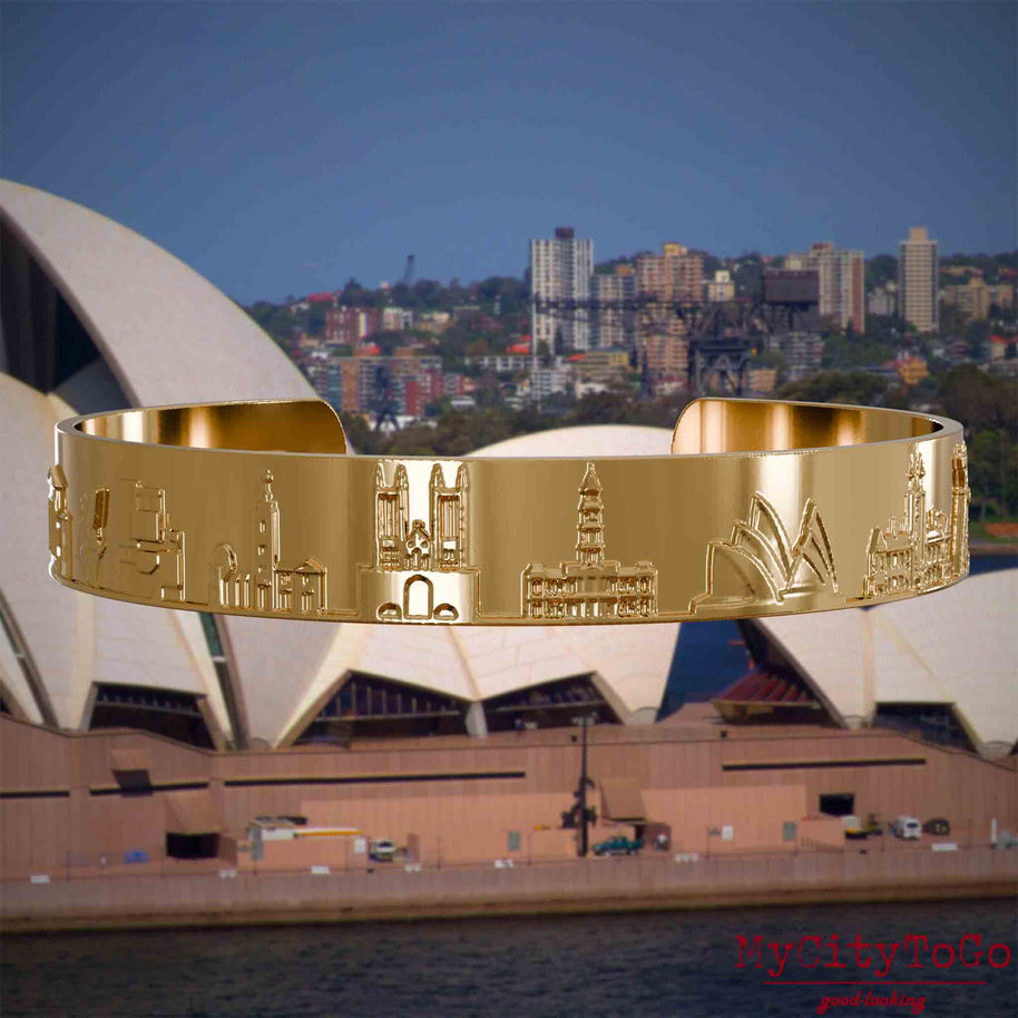 Vergoldeter Armreif mit Reliefs bekannter Sehenswürdigkeiten der Stadt Sydney. Den Hintergrund bildet eine Ansicht der Stadt Sydney.