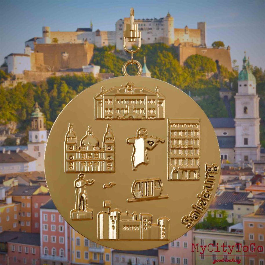 Halskette mit Drehwirbel und Vorderseite eines vergoldeten Schmuckanhängers, auf dem Reliefs bekannter Sehenswürdigkeiten der Stadt Salzburg abgebildet sind. Den Hintergrund bildet eine Ansicht der Stadt Salzburg.
