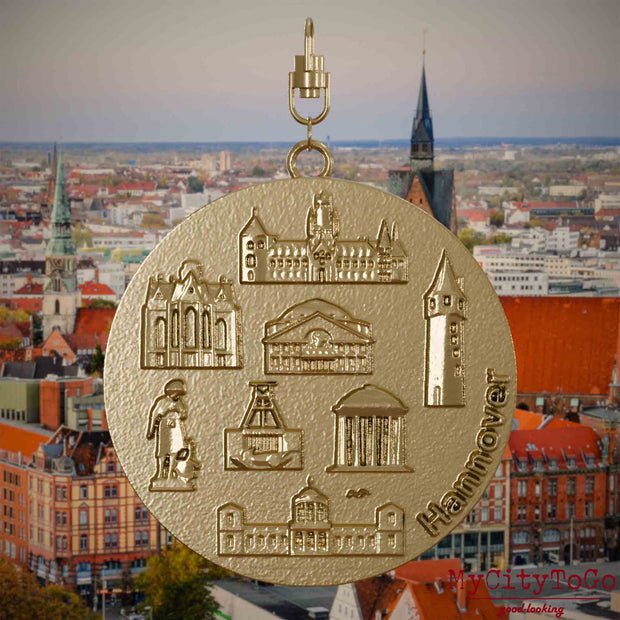 Halskette mit Drehwirbel und Vorderseite eines Schmuckanhängers aus vergoldetem Edelstahl, auf dem Reliefs bekannter Sehenswürdigkeiten der Stadt Hannover abgebildet sind. Den Hintergrund bildet eine Ansicht der Stadt Hannover .
