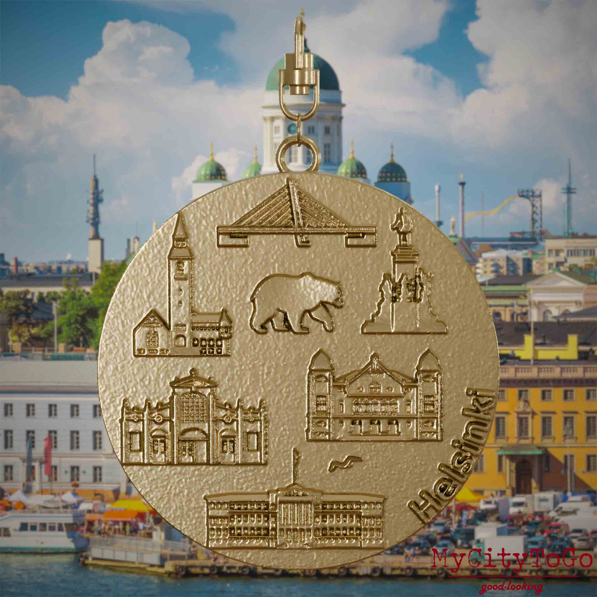 Halskette mit Drehwirbel und Rückseite eines Schmuckanhängers aus vergoldetem Edelstahl, auf dem Reliefs bekannter Sehenswürdigkeiten der Stadt Helsinki abgebildet sind. Den Hintergrund bildet eine Ansicht der Stadt Helsinki.
