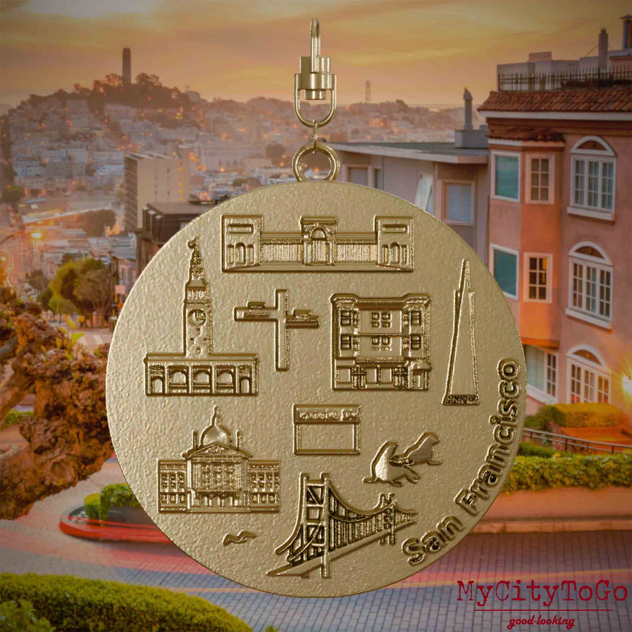 Halskette mit Drehwirbel und Vorderseite eines Schmuckanhängers aus vergoldetem Edelstahl, auf dem Reliefs bekannter Sehenswürdigkeiten der Stadt San Francisco abgebildet sind. Den Hintergrund bildet eine Ansicht der Stadt San Francisco.
