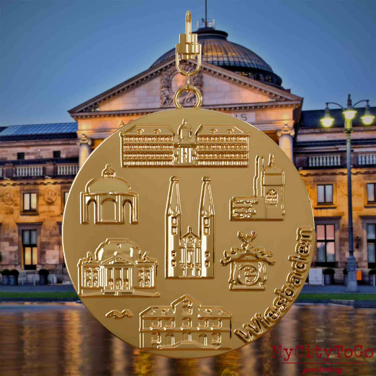 Halskette mit Drehwirbel und Rückseite eines vergoldeten Schmuckanhängers, auf dem Reliefs bekannter Sehenswürdigkeiten der Stadt Wiesbaden abgebildet sind. Den Hintergrund bildet eine Ansicht der Stadt Wiesbaden.
