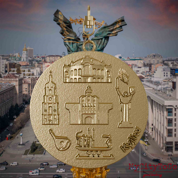 Halskette mit Drehwirbel und Vorderseite eines Schmuckanhängers aus vergoldetem Edelstahl, auf dem Reliefs bekannter Sehenswürdigkeiten der Stadt Kyjiw abgebildet sind. Den Hintergrund bildet eine Ansicht der Stadt Kyjiw.
